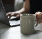 Photo of a woman holding a coffee mug and working on a laptop.